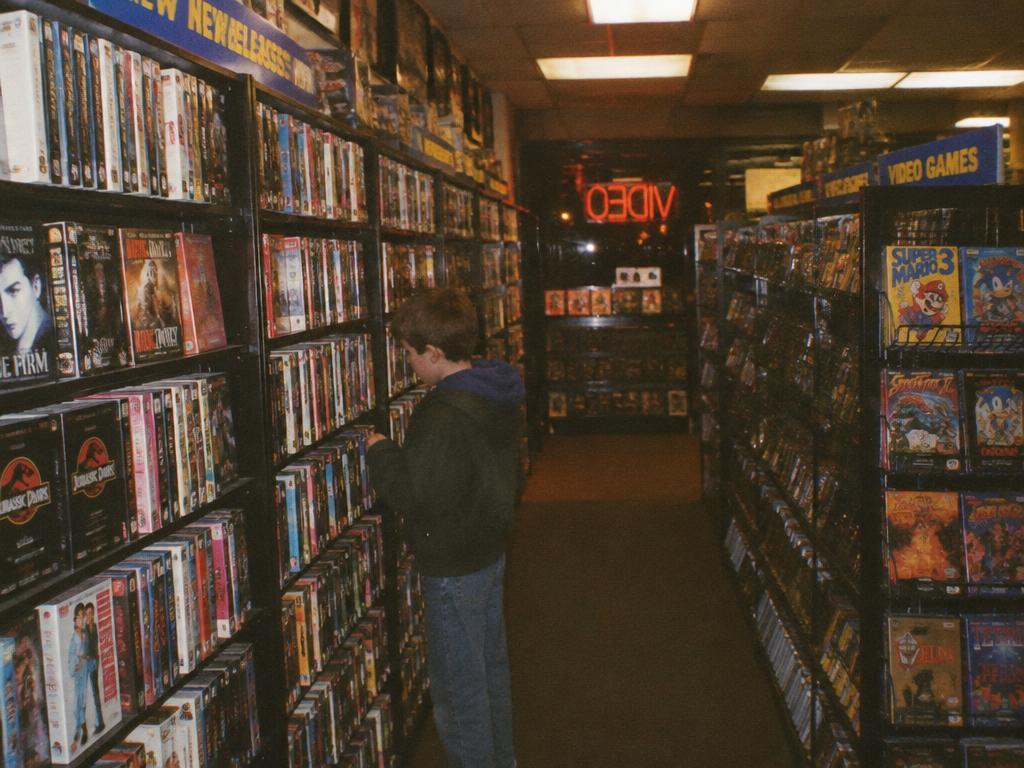 A kid browsing a 1990s video rental aisle lined with tapes and game boxes